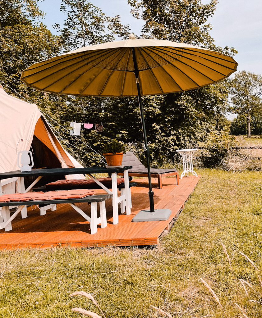 De glamping tent in Vorden, met op de voorgrond een ruime picknicktafel in de buitenlucht. Een parasol biedt schaduw tegen de felle zonnestralen die door de bomen schijnen. De tent op de achtergrond is ruim en comfortabel ingericht met een groot tweepersoonsbed en zachte kussens en dekens. De omgeving is groen en bosrijk, met een heldere blauwe lucht op de achtergrond.