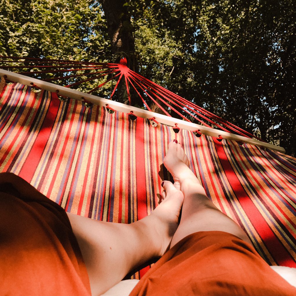 Een vrouw die ligt te relaxen in een hangmat met de voeten omhoog in de zon. De hangmat is bevestigd tussen twee bomen, vlakbij een glamping tent op het terrein van B&B Erve Hasselo in Vorden. Op de achtergrond zijn bomen te zien. De sfeer straalt rust en ontspanning uit.