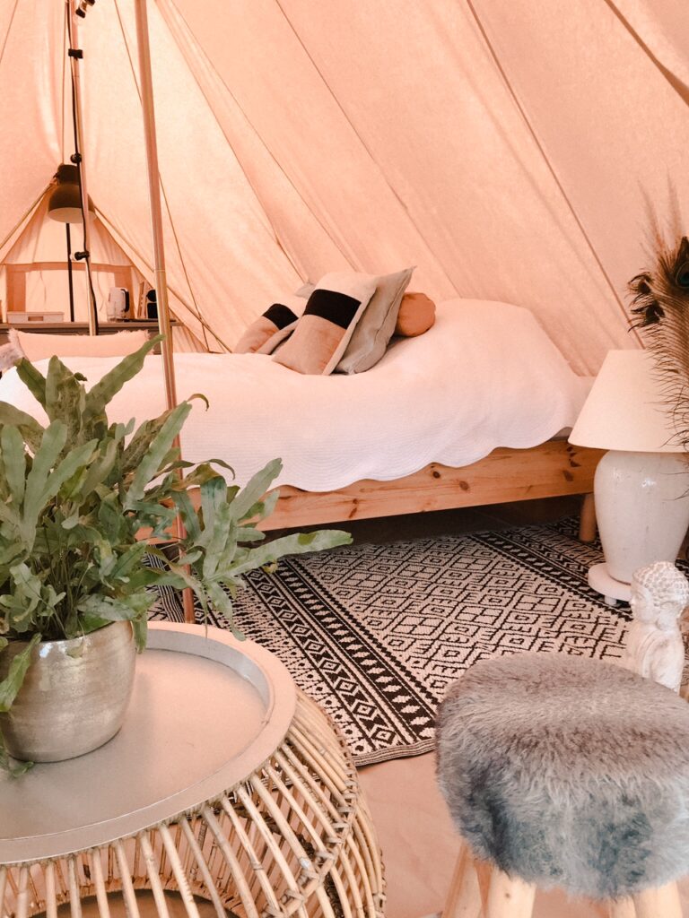 Een riant bed met wit linnen beddengoed en kussenns, in de glamping tent bij Erve Hasselo in Vorden. Op de voorgrond een tafeltje met een plant en een kruk.