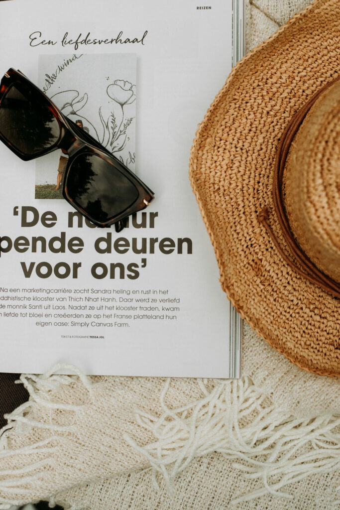 Zonnebril en zonnehoed liggend op een opengeslagen tijdschrift en een crème kleurig plaid.